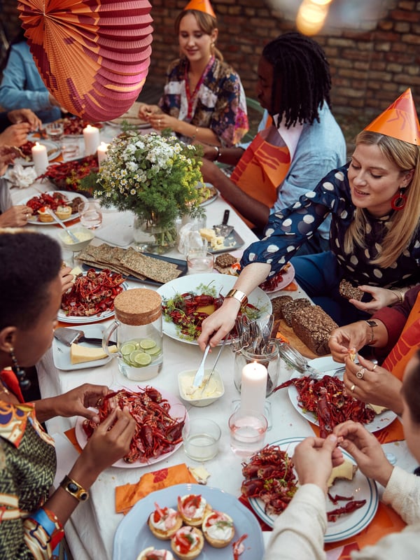 Grupp människor runt ett bord med kräftor, sallad, bröd och dryck. Ljus och blommor dekorerar bordet. Några bär partyhattar.
