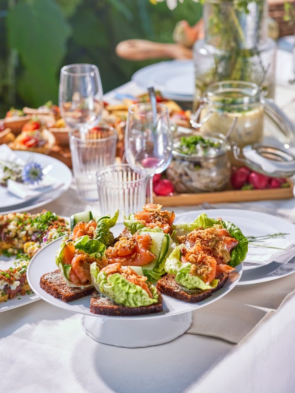 A serving stand with crispy bread sandwiches placed on a table outdoors, with other food served on plates and chopping boards.