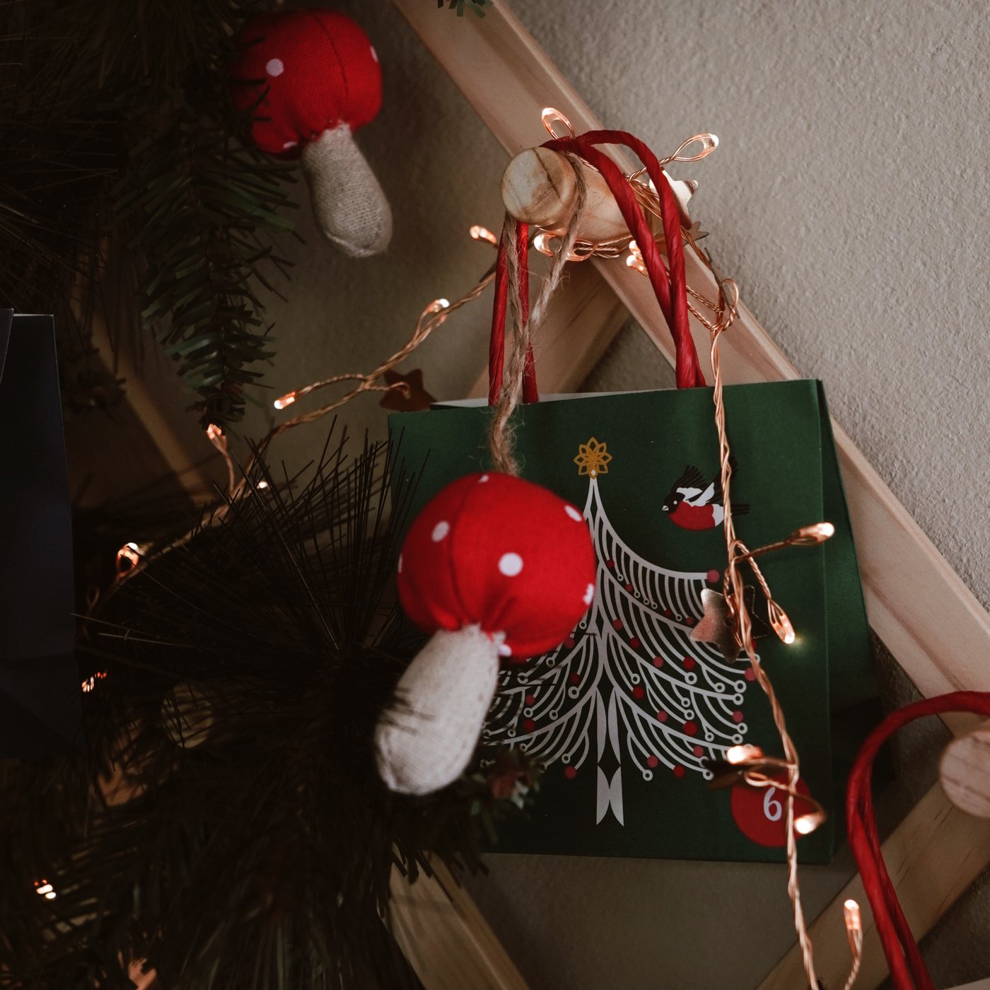 Weihnachtliche Szene mit roten Pilzornamenten und Lichterketten an einem Baum. Im Hintergrund steht eine grüne Geschenktüte mit einem weißen Baummotiv.