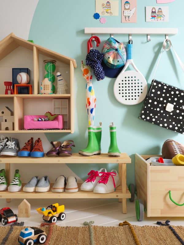 Una rastrelliera per scarpe sul pavimento sotto a una casa delle bambole/scaffale FLISAT montata a parete con accanto un contenitore per giocattoli con rotelle - IKEA.