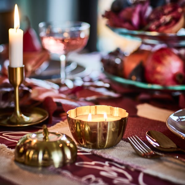 A lit HÖSTAGILLE scented candle in a pumpkin-shaped metal tin is on a dining table with a pink and red autumn theme.