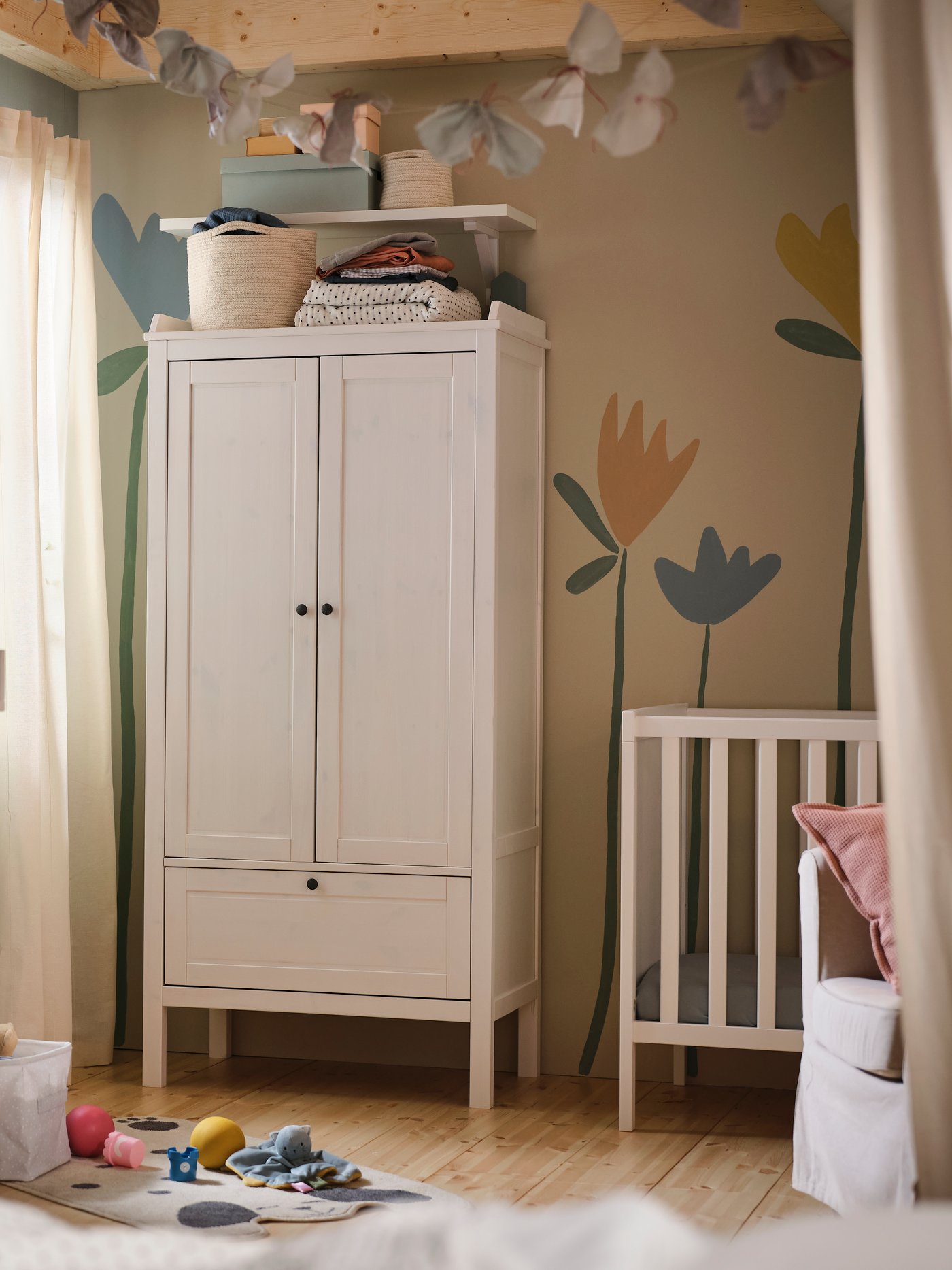 In einem Babyzimmer steht ein weisser SUNDVIK Kleiderschrank an einer Wand mit einem Blumen-Wandbild. Daneben befindet sich ein weisses SUNDVIK Babybett.