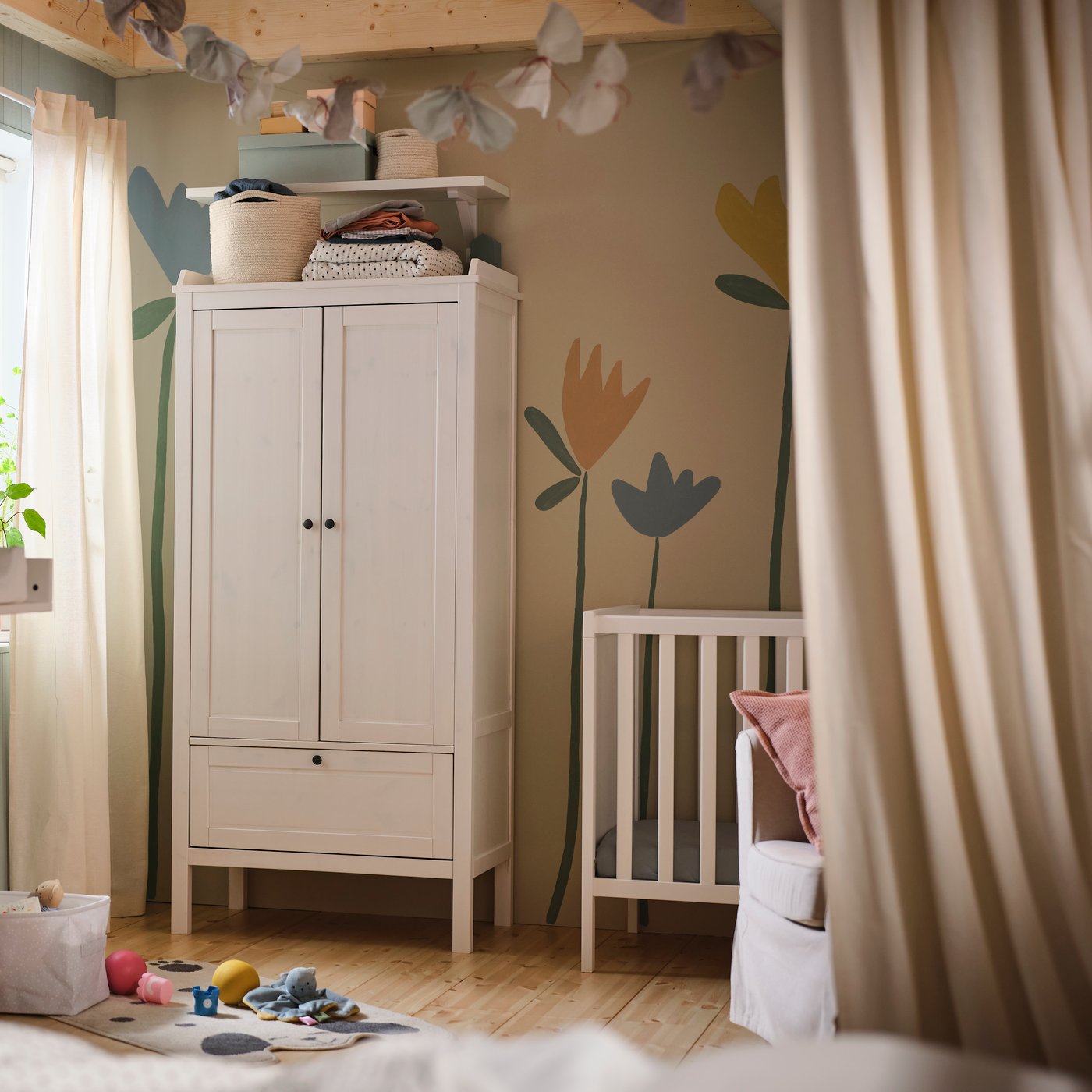 A white SUNDVIK wardrobe against a wall with a flower mural on it in a baby’s bedroom with a white SUNDVIK cot beside it.