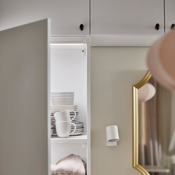 A white PLATSA wardrobe, an open door revealing tableware inside. Next to it hangs a SVANSELE gold-colour mirror.