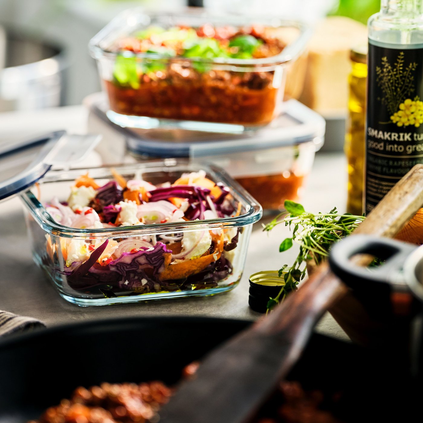 A IKEA 365+ glass food storage container with prepared food is next to a pan. Two other containers are stacked behind it.