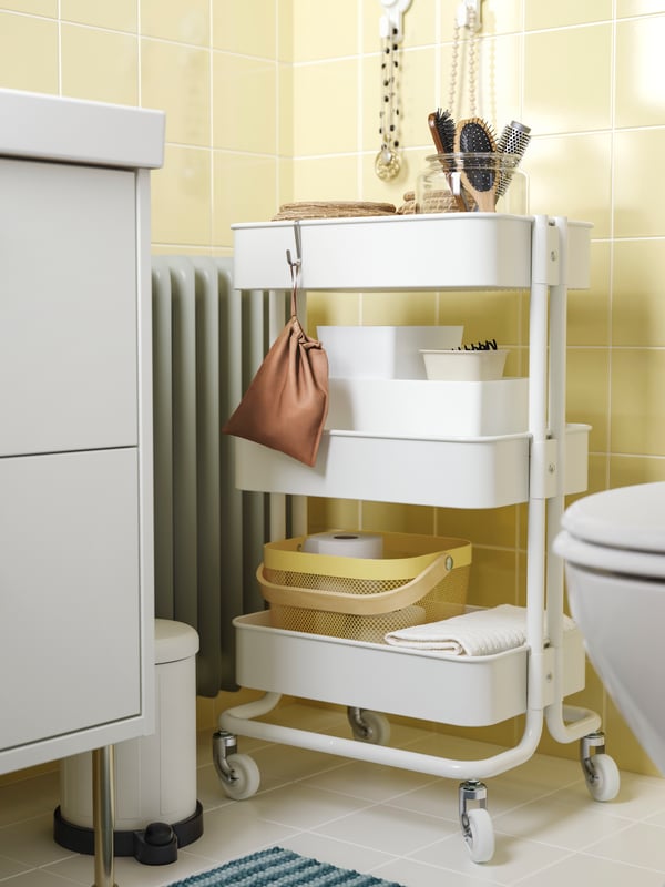 A white RÅSKOG trolley, with bathroom items stored inside, is placed in a corner, between a toilet and a HAVBÄCK washstand.