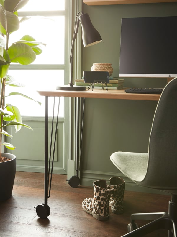A dimly sunlit workspace with a table on castors, a LÅNGFJÄLL conference chair, a big nearby plant and leopard-pattern shoes.