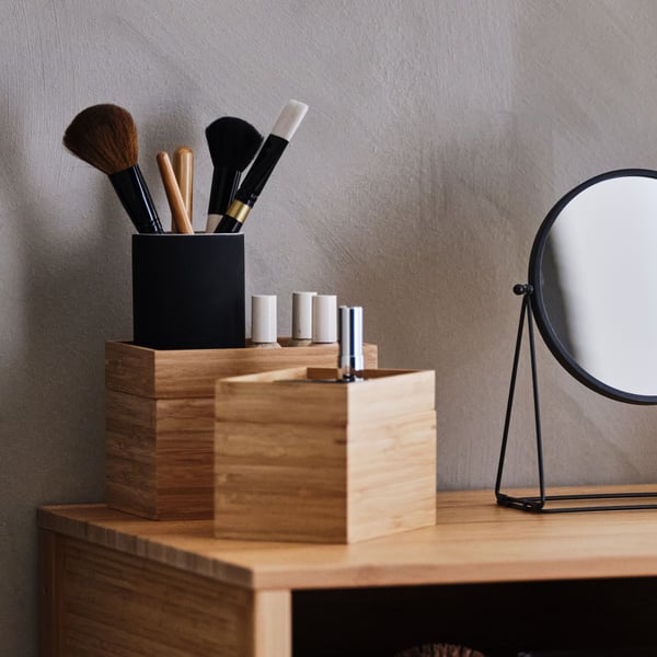 Two containers made of bamboo with cosmetics and makeup brushes are standing on a wooden shelf next to a table mirror.