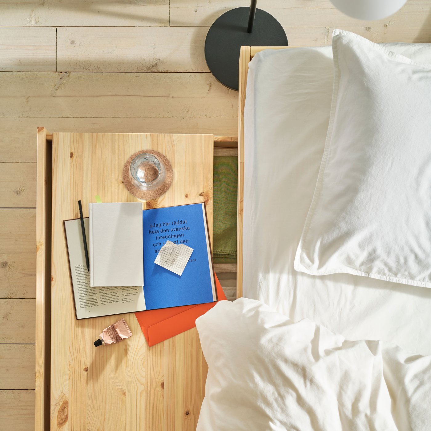 Shot from above on bed with white sheets, drawer below pulled out with tray in natural wood, a book and a glass on tray.