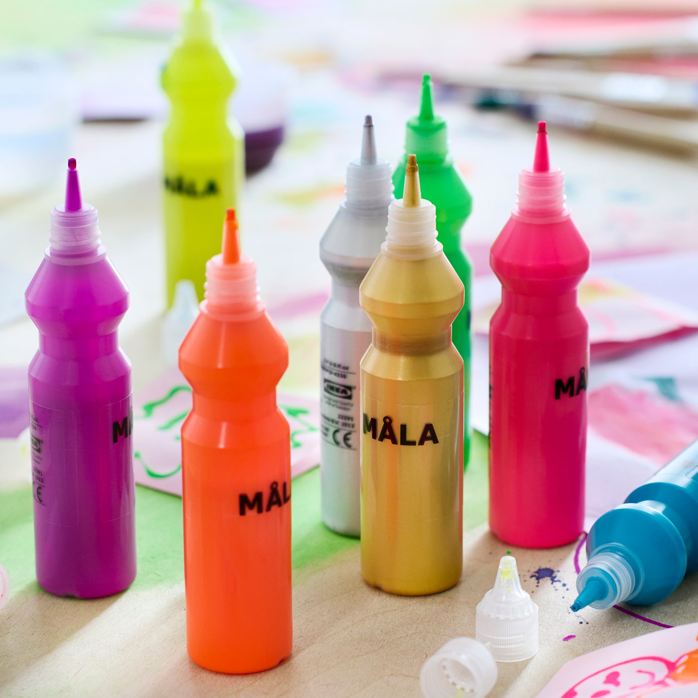 Gros plan de huit bouteilles de peinture fluorescente MÅLA entourées de peintures et de dessins variés sur une table.
