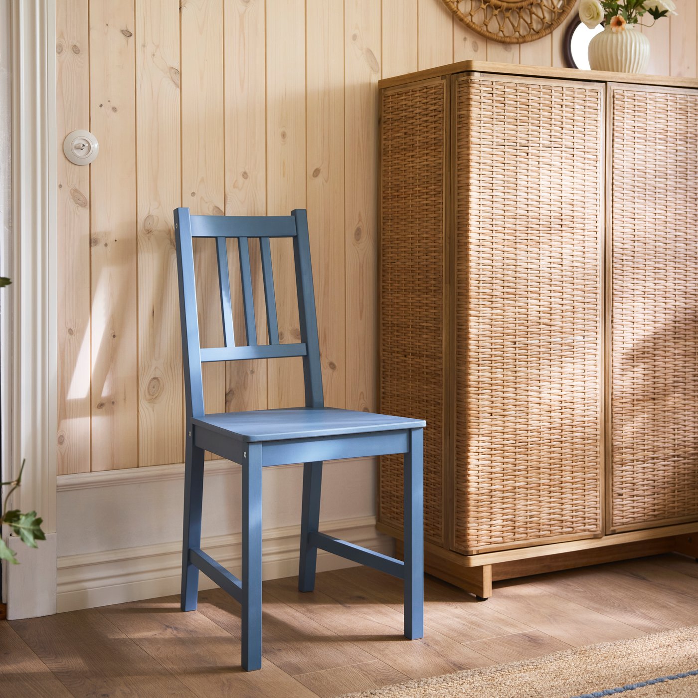 Blue PINNTORP chair standing in a light wooden room next to a woven cabinet.