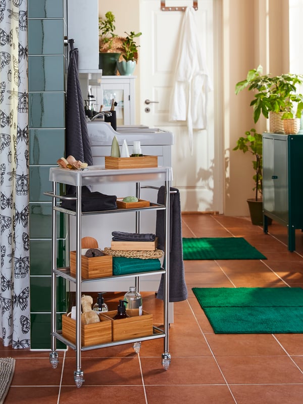 A GRUNDTAL trolley in stainless steel is standing next to a shower curtain, the shelves stacked with beauty products.