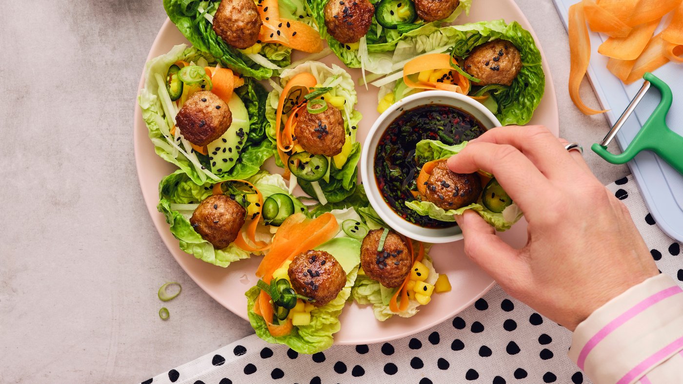 Eine Person hält ein in ein Salatblatt eingeschlagenes Fleischbällchen und taucht es in eine weisse Schüssel mit Sojasosse.