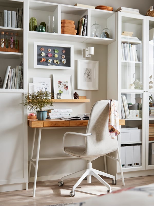An IKEA LÅNGFJÄLL conference chair in beige with white legs and armrests in a sunny office with white bookcases and a wooden desk with white legs.