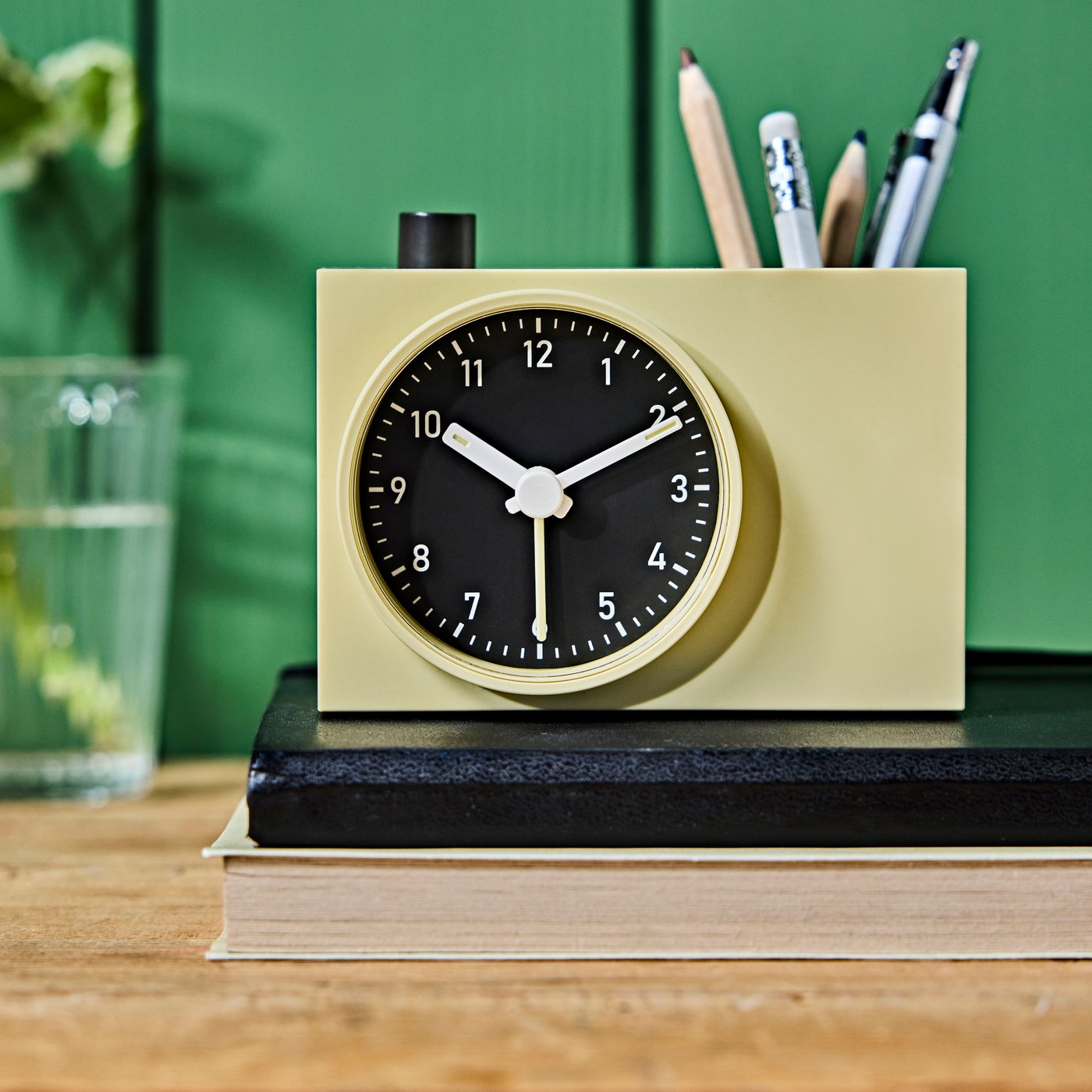 A close-up of a yellow ÖKENMARSVIN analogue alarm clock, holding pens and pencils, placed on two books lying on a table.
