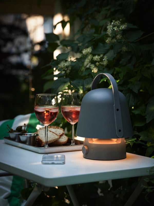 VAPPEBY Bluetooth zvučnik-lampa nalazi se na stolu, pored tacna za serviranje na kojoj su dve vinske čaše i grickalice.