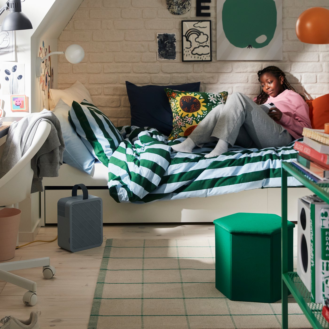A teenage girl is relaxing on a white bed dressed with a green‑blue striped duvet cover in a teen room with colourful furniture and accessories.