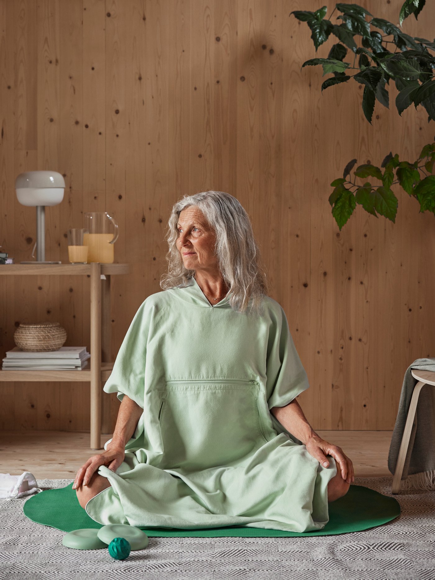 A woman sits cross-legged on a small DAJLIEN exercise mat in a living room, wearing the DAJLIEN bath poncho with a hood.