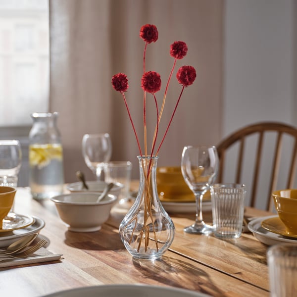 Clear glass bud vase with decorative red dried stems displayed as a dining table centerpiece in a warm, sunlit setting.