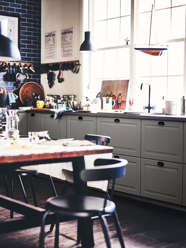 A kitchen with grey BODBYN drawer fronts, a worktop, a sink, a table, YNGVAR chairs and a dark grey HEKTAR pendant lamp.