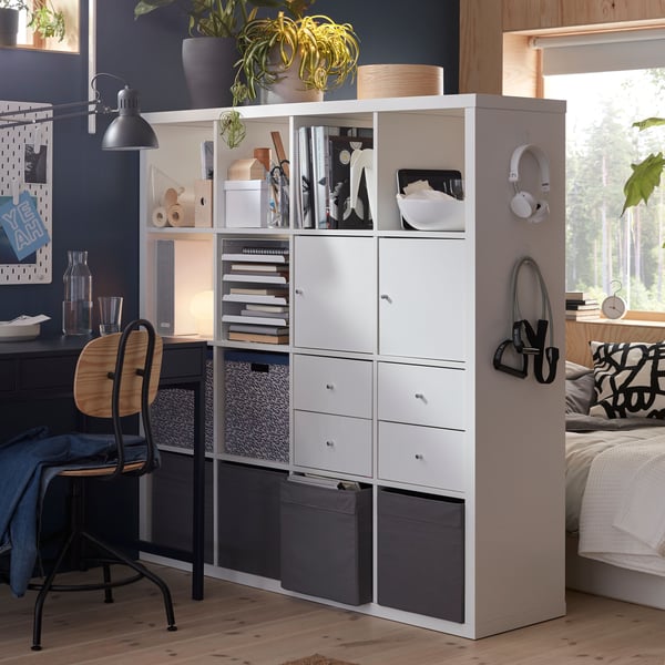Een kamer verdeeld met een witte KALLAX-rek met vier inzetstukken en DRÖNA-boxen, waardoor een slaapgedeelte en een werkruimte ontstaan.