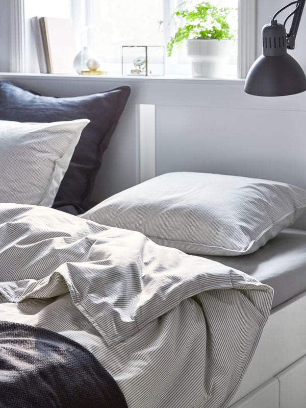 A white NORDLI bed frame with storage underneath. Two of the drawers are open and contain books, a bedspread and various items.