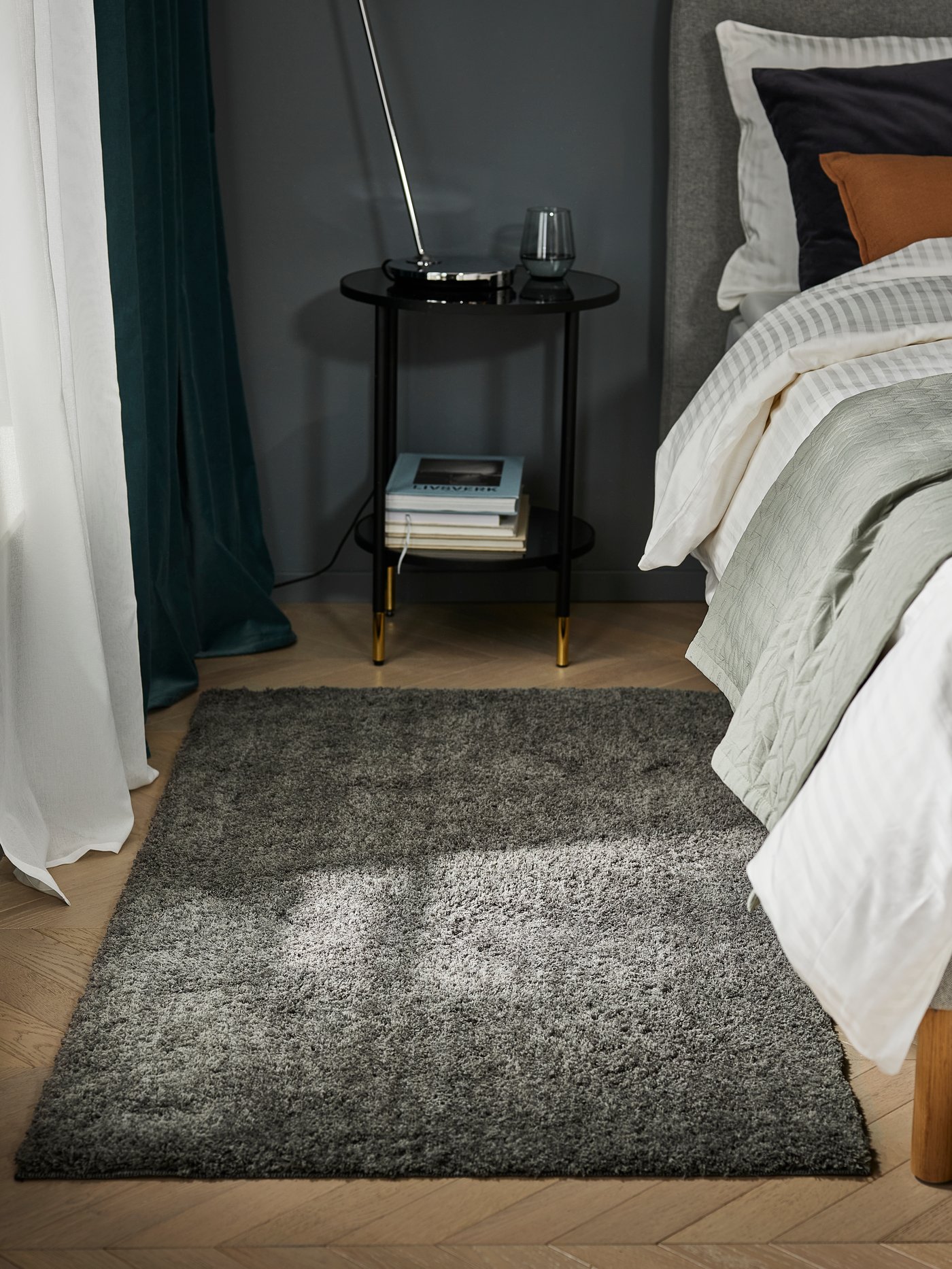 Close-up of LINDKNUT rug in dark grey on floor between window and bed, with ÄSPERÖD side table in back.