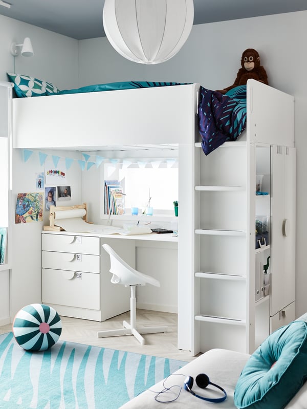 A white loft bed with torquoise bedding, a desk and swivel chair below with crafting materials and blue bunting, a blue rug.