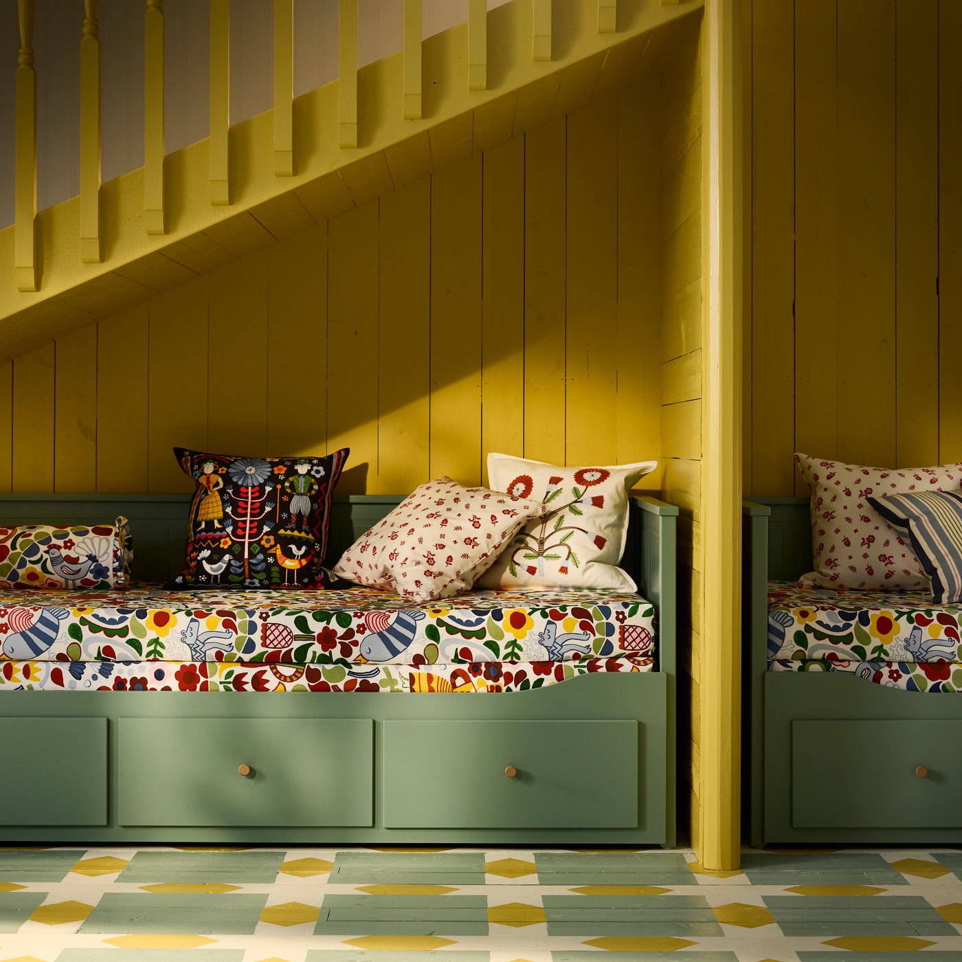 Two grey-green HEMNES day-bed frames with 3 drawers sit against a yellow wall. There are several colourful cushions on them.