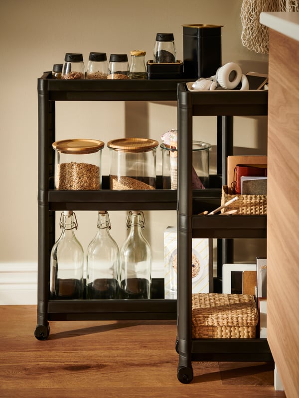 A kitchen corner with two side-by-side black NISSAFORS trolleys, one holding food, jars and bottles, the other paperwork.