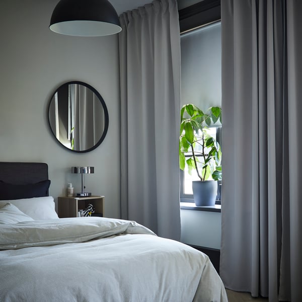 Soft light falls on a bed from a window with open MAJGULL block-out curtains in light grey turquoise over sheer grey curtains.