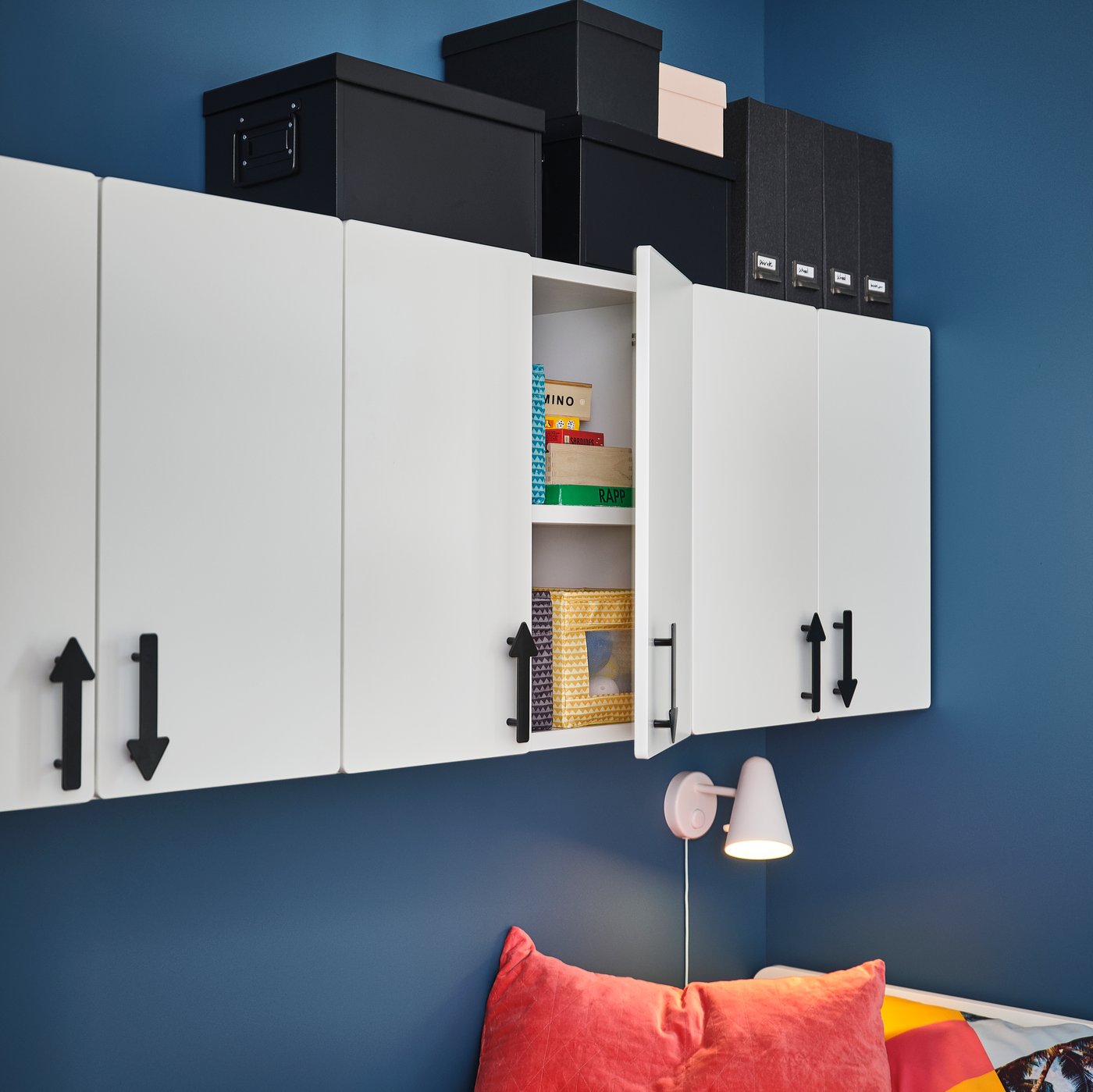 Three white SMÅSTAD wall cabinets on a blue wall with the door of the middle cabinet slightly open showing games inside.