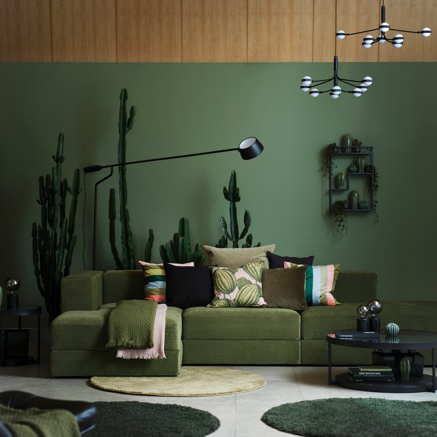 In einem Wohnzimmer ist ein JÄTTEBO 3,5er-Sitzelement mit Récamieren mit Bezug „Samsala“ in Dunkelgelbgrün, gemusterten Kissen und Plaids zu sehen. Hinter dem Sofa befinden sich viele Kakteen.