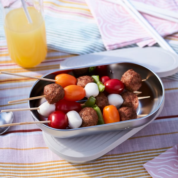 Envase para guardar comida metálico HALVARM sobre una manta de picnic. Enel interior del envase  hay brochetas con albóndigas IKEA, tomates cherry y otros vegetales. 