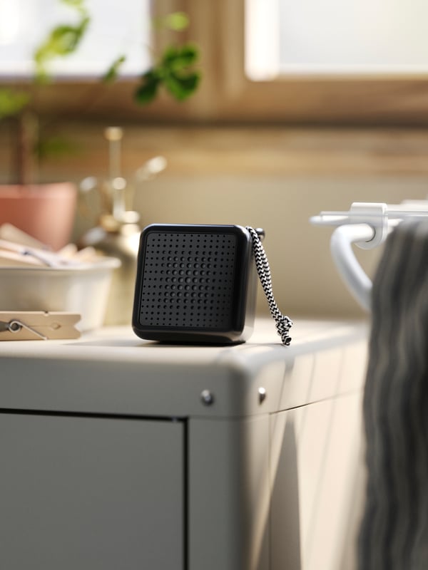 سماعة صغيرة VAPPEBY سوداء يمكن حملها ومقاومة للماء تعمل بتقنية Bluetooth بالقرب من وعاء مشابك أعلى خزانة KOLBJÖRN.