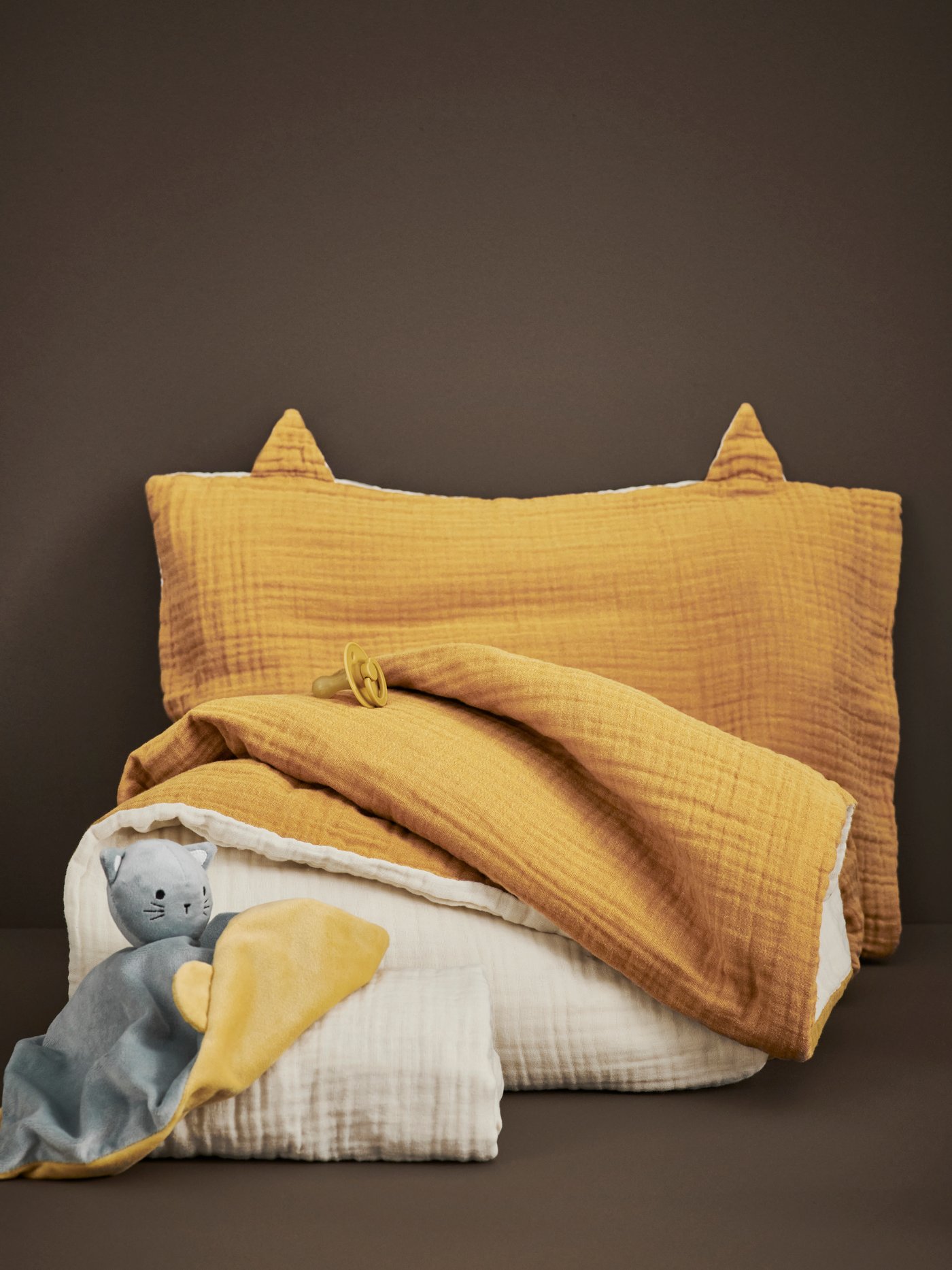 Close up of GULDVÄVARE 3-piece bedlinen set on a brown floor. Next to it there’s a GULDVÄVARE comfort blanket with soft toy.