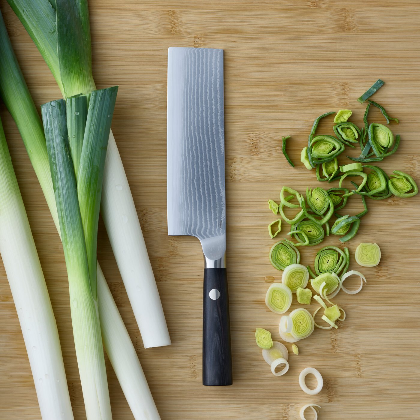 A close-up of a black FJÄLLRÖDING nakiri knife made of Damascus steel, placed next to freshly sliced leeks on a cutting board.