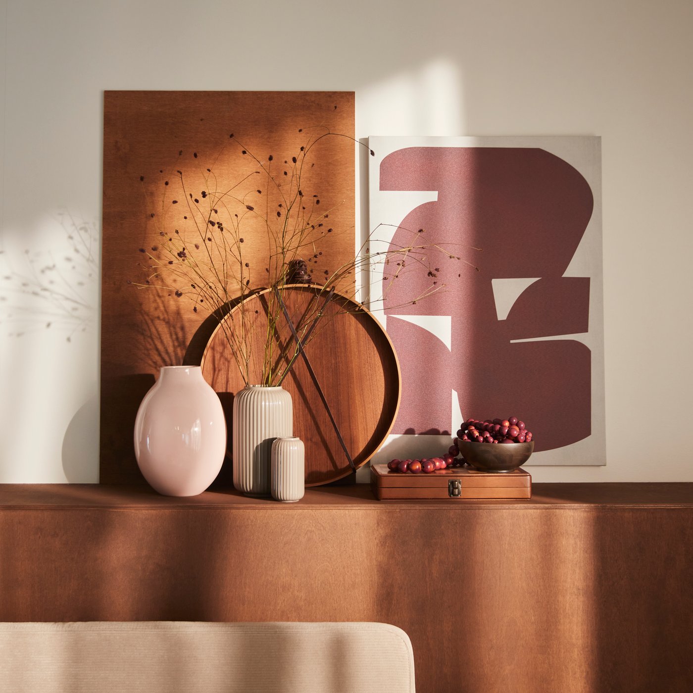 Auf einer hellbraunen Fläche stehen eine blassrosa BLODBJÖRK-Vase, weitere Vasen, Dekoartikel und ein PJÄTTERYD-Bild.