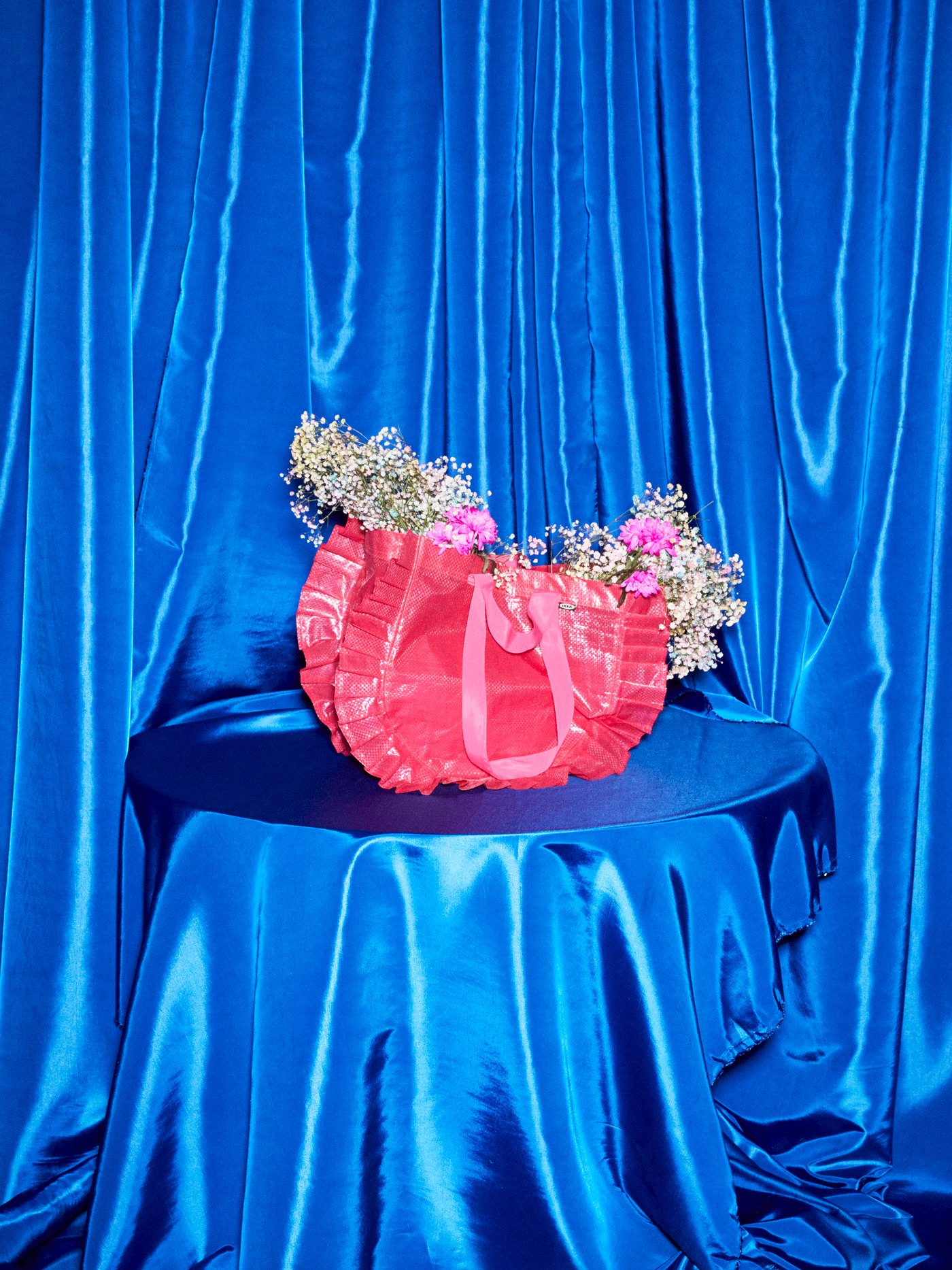 A medium KARISMATISK pink carrier bag sits atop a table completely covered in blue satin; the bag is filled with flowers.