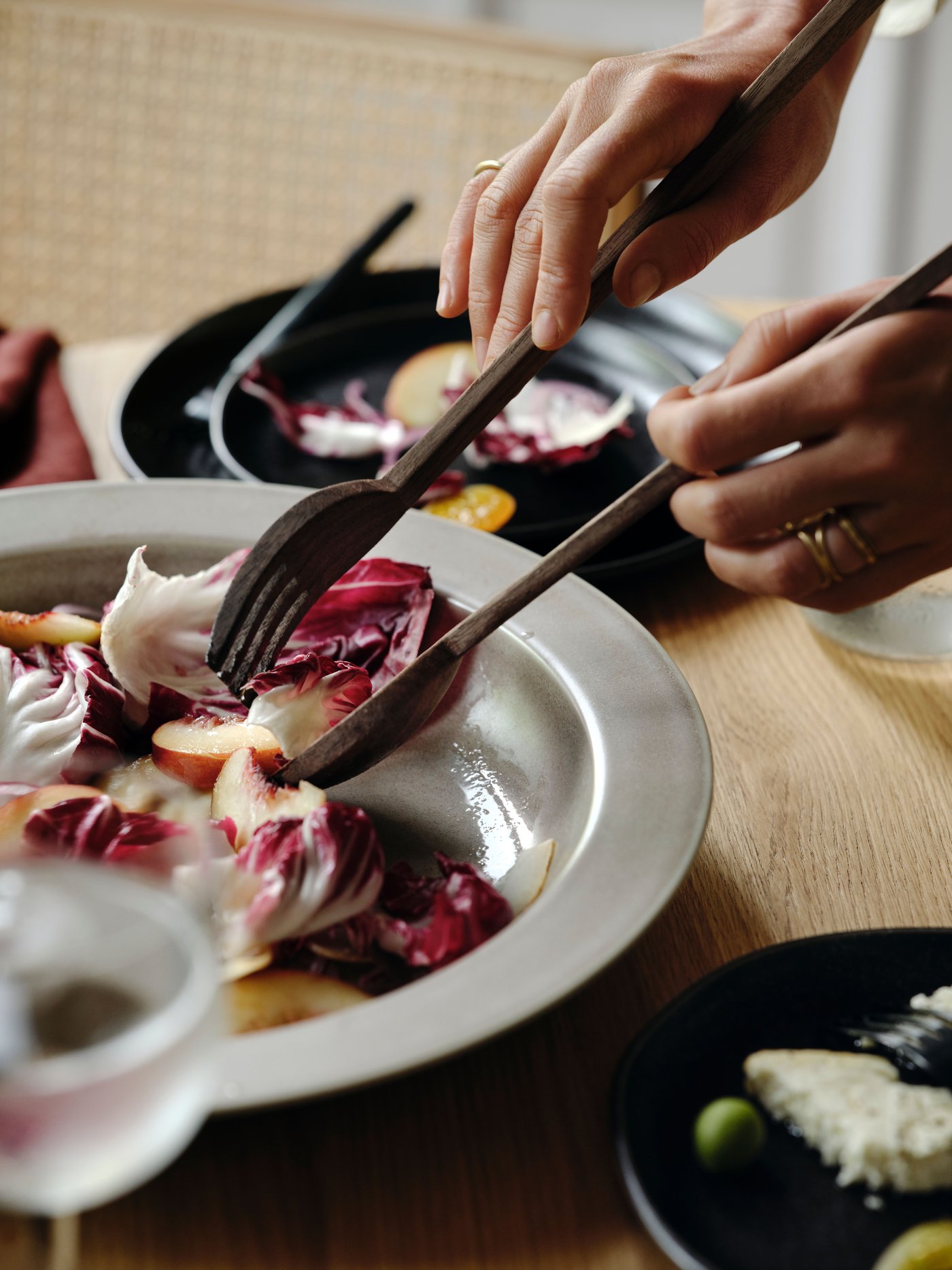 Två händer som håller ett STOCKHOLM 2025 serveringsset i två delar i valnöt och plockar mat ur en beige serveringsskål.
