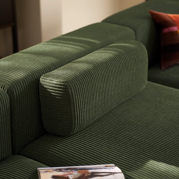 A loose, matching armrest placed by the backrest of a dark-green JÄTTEBO four-seat sofa, flanked by a cushion and a magazine.