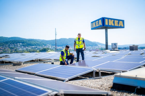 Deux techniciens en gilets de sécurité inspectent des panneaux solaires sur le toit d’un bâtiment IKEA, collines en arrière-plan.