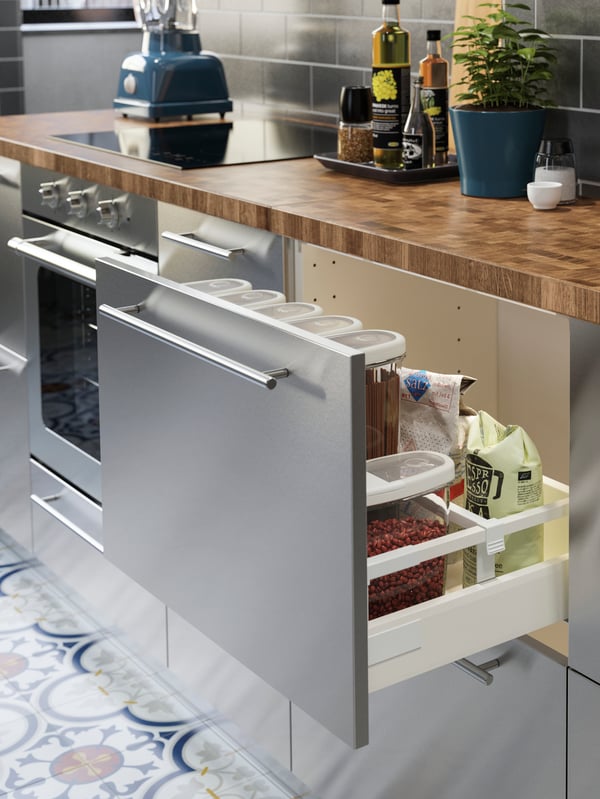 A stainless steel kitchen with a wooden worktop in oak/veneer. One drawer is open, dry goods are stored inside.