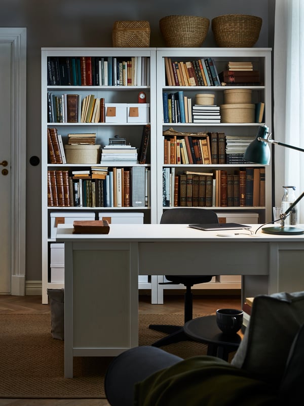 A workspace at home with a white HEMNES desk and matching bookcases placed behind it with books and boxes.
