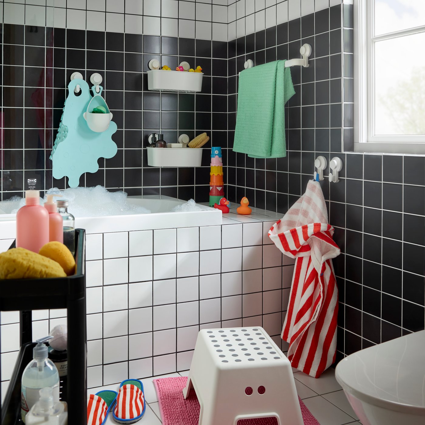 A black-and-white bathroom with a white BOLMEN step stool on a pink bath mat. A red striped poncho, slippers and toys.