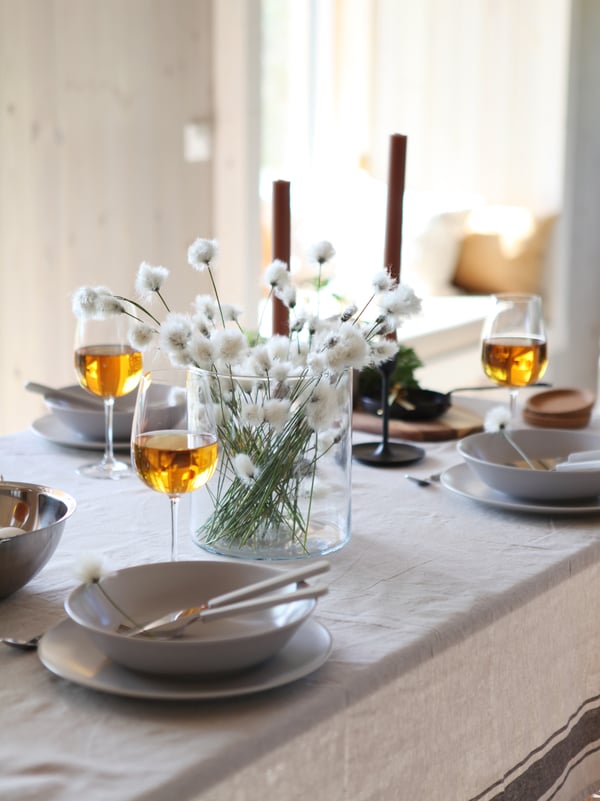 An IKEA set of FÄRGKLAR dinnerware in light grey set up on a light grey tablecloth. Wine glasses on the table, a clear vase set with white flowers and black candlesticks. 