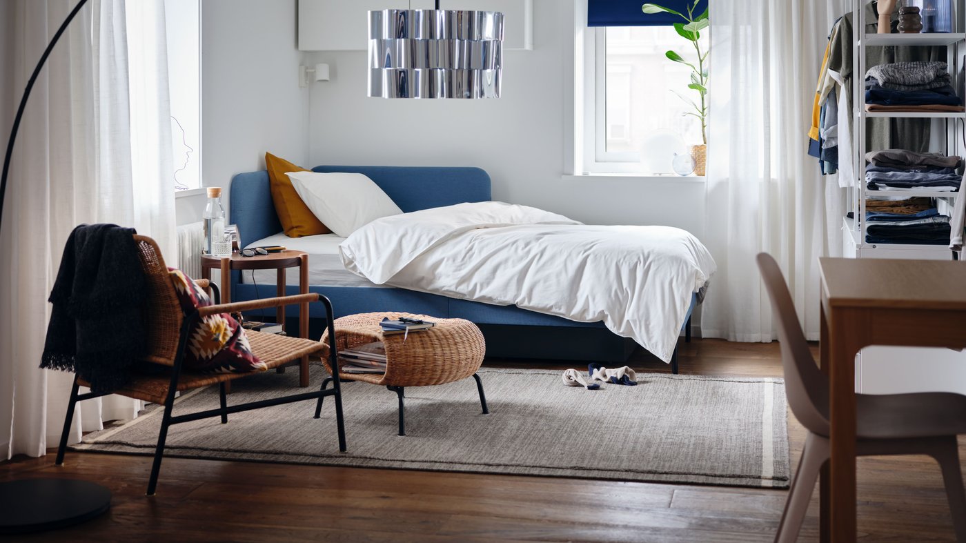 Un dormitorio con una estructura de cama tapizada con respaldo de esquina, funda de edredón y funda de almohada blancas y una planta en maceta.
