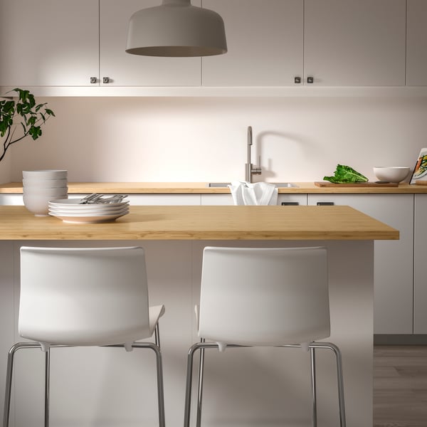 A kitchen with HOLMARED custom-made worktop in bamboo/veneer on the kitchen island, tableware, and two white kitchen chairs. 