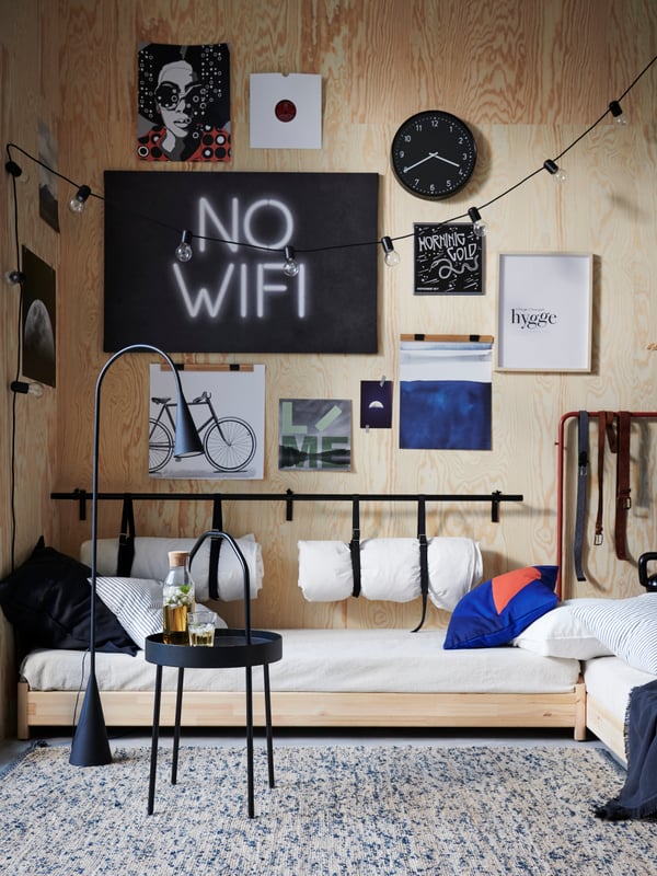 Two rolled-up duvets hang from a RÄCKA curtain rod on a plywood wall, above one section of an UTÅKER stackable bed.