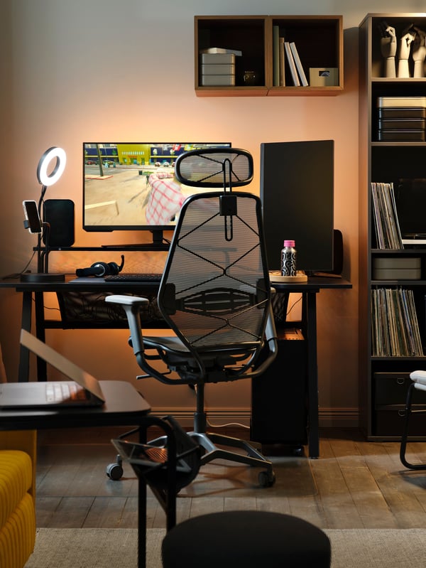 A glowing, backlit PC gaming setup has a paused game on a monitor on a black UTESPELARE gaming desk by a grey mesh chair.